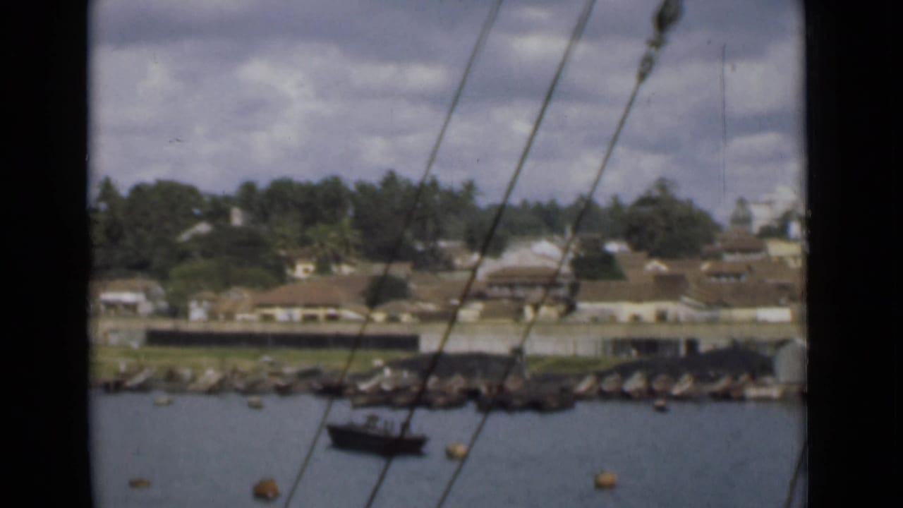 View across Colombo harbor water toward shoreline buildings, trees, rocks, and a small boat, seen through vessel rigging lines.