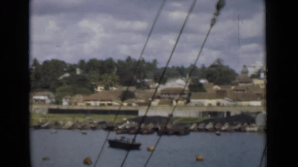View across Colombo harbor water toward shoreline buildings, trees, rocks, and a small boat, seen through vessel rigging lines.