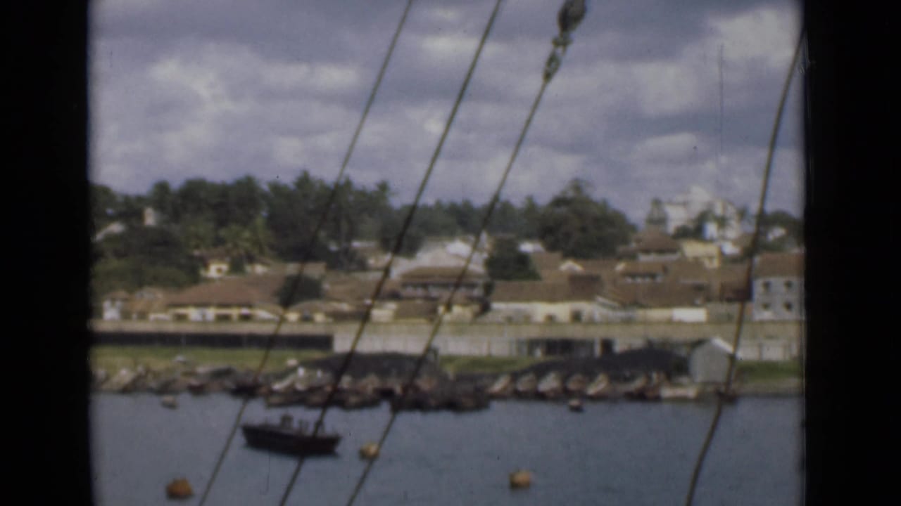 Colombo harbor shoreline appears beyond water and rigging, with a small boat near rocks and low buildings among trees.