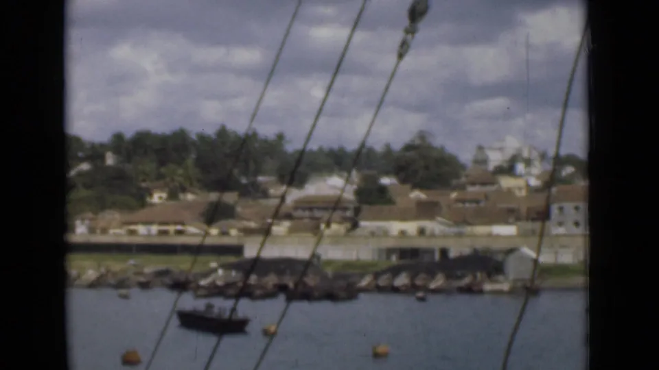 Colombo harbor shoreline appears beyond water and rigging, with a small boat near rocks and low buildings among trees.