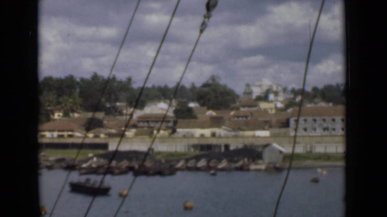 Steady 1946 home-movie view across Colombo harbor, with rigging lines in front of a small boat, rocky shore, trees, and waterfront buildings.
