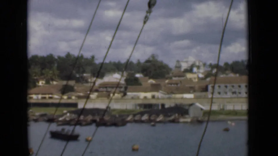 Steady 1946 home-movie view across Colombo harbor, with rigging lines in front of a small boat, rocky shore, trees, and waterfront buildings.