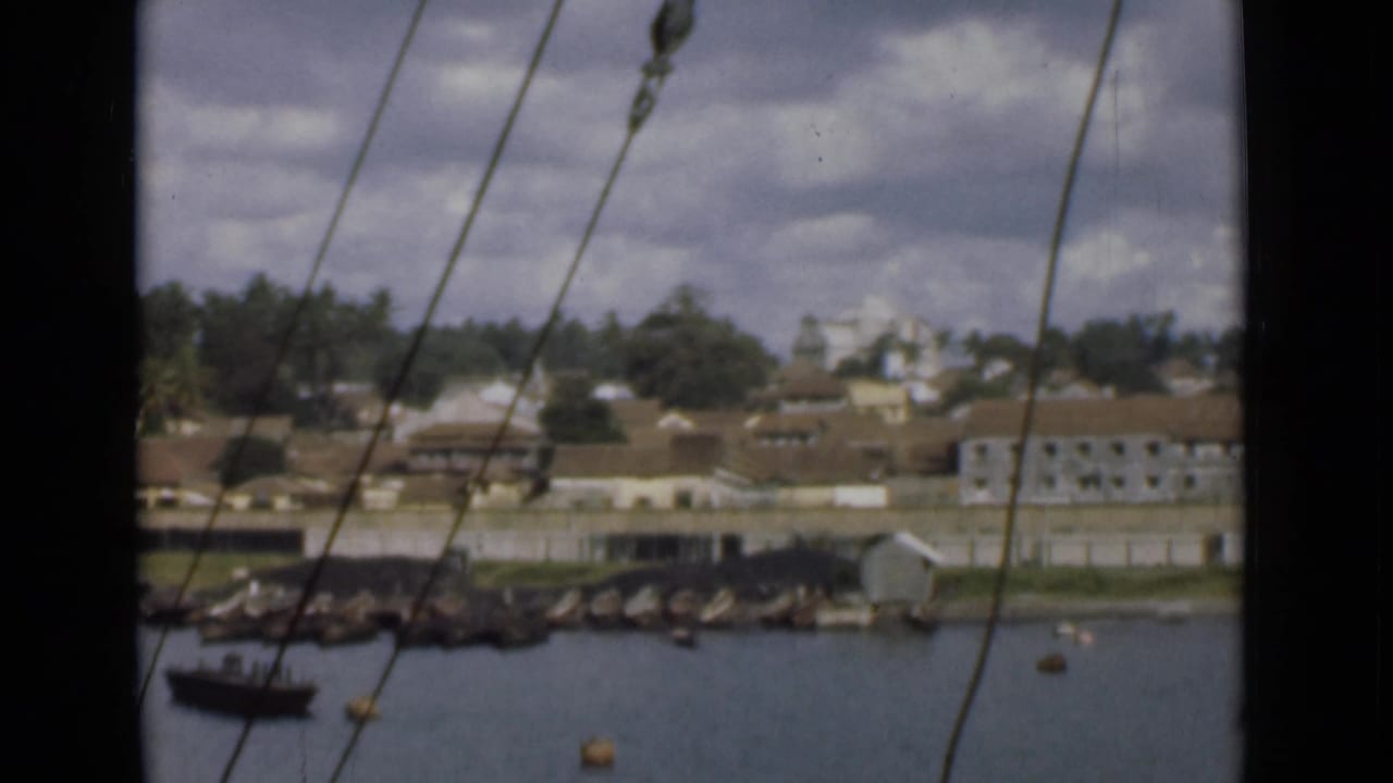 1946 Colombo harbor view from a ship, with rigging crossing the frame and low waterfront buildings beyond the water.