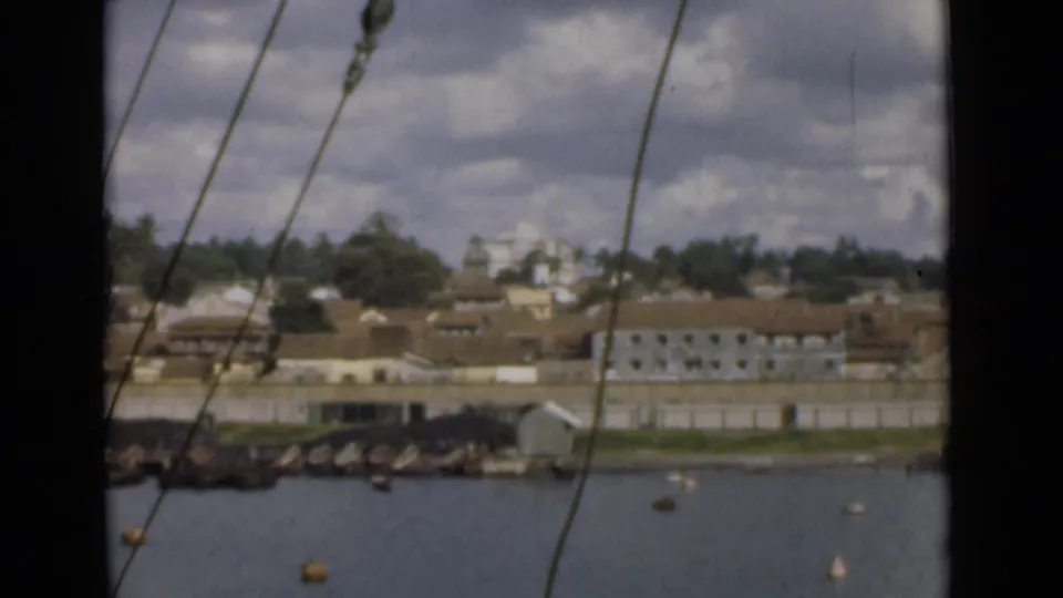Shipboard 8mm view of Colombo waterfront buildings across harbor water, partly framed by rigging lines.