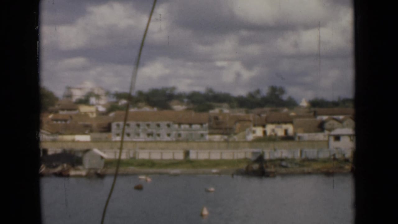 1946 Colombo, Ceylon waterfront seen across calm water, with low buildings, trees, buoys, and cloudy sky in a soft 8mm frame.
