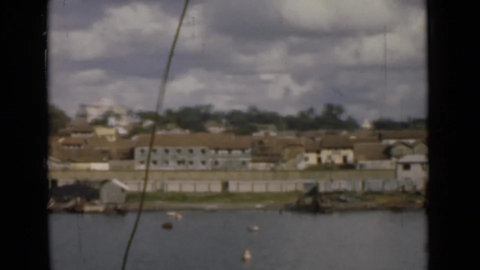 1946 Colombo, Ceylon waterfront seen across calm water, with low buildings, trees, buoys, and cloudy sky in a soft 8mm frame.