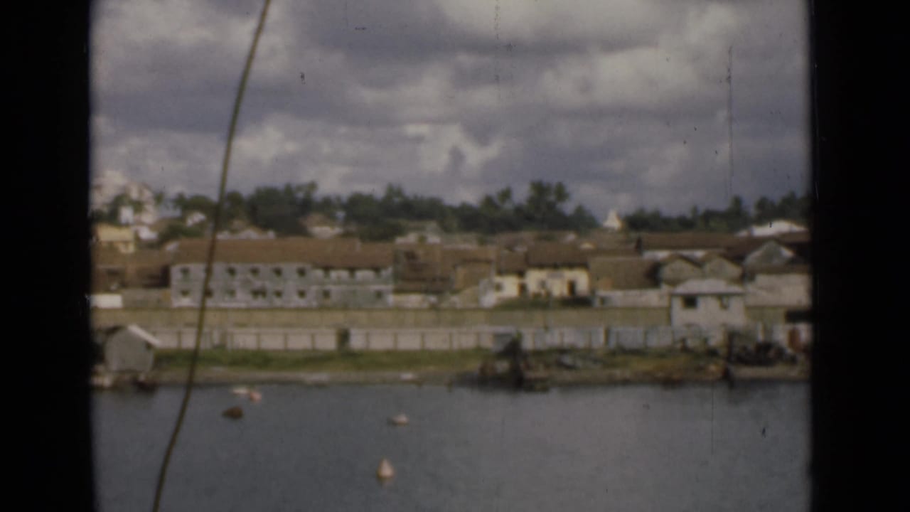 Colombo shoreline from across the water, showing low-rise buildings, a seawall, trees, buoys, and overcast sky on 8mm film.