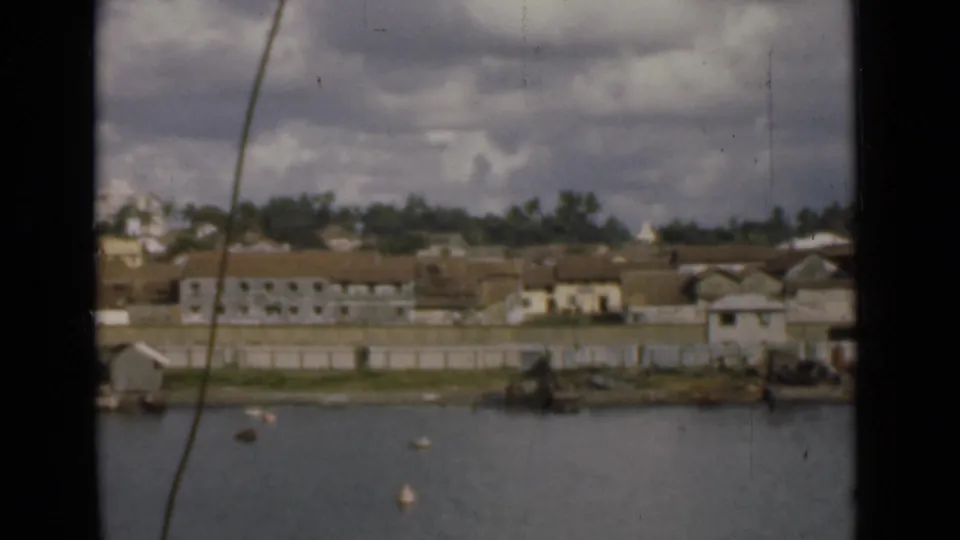 Colombo shoreline from across the water, showing low-rise buildings, a seawall, trees, buoys, and overcast sky on 8mm film.