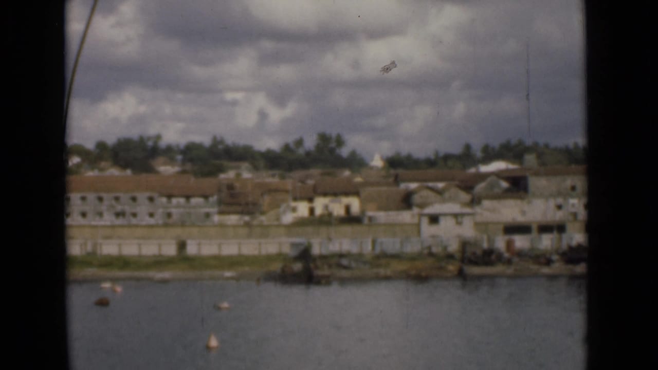 Mid-1940s Colombo waterfront view with tiled-roof buildings, a pale seawall, trees, and floating markers under cloudy light.