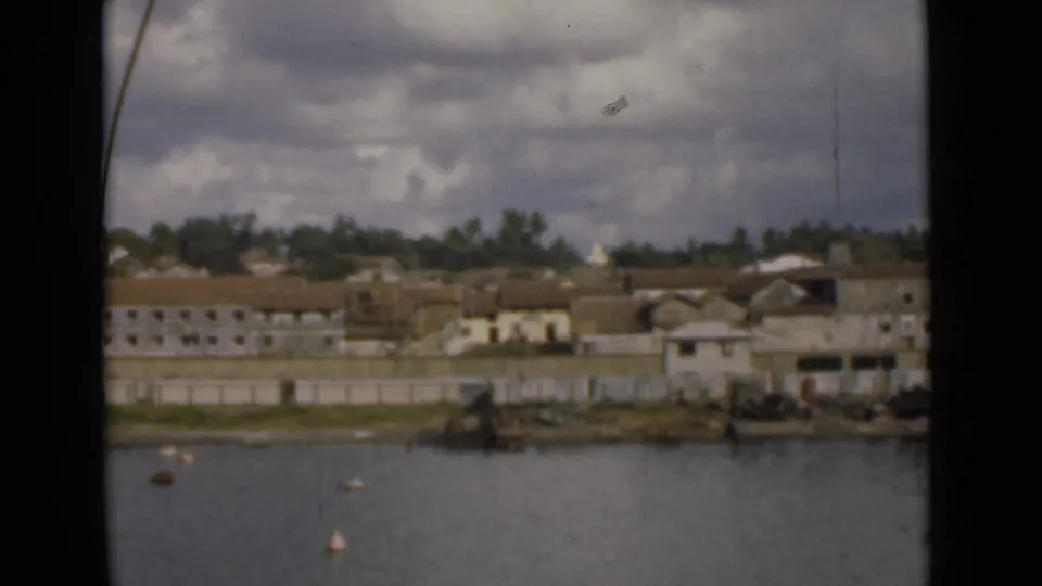 Mid-1940s Colombo waterfront view with tiled-roof buildings, a pale seawall, trees, and floating markers under cloudy light.