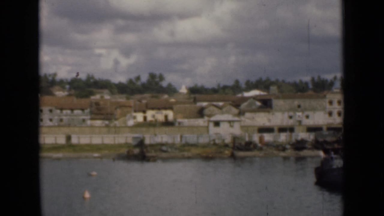 Distant Colombo waterfront on 8mm film, with calm water, buoys, low buildings, green trees, clouds, and a dark frame edge.