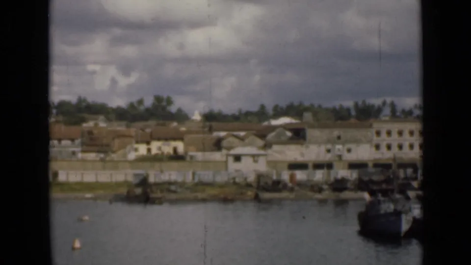 1940s Colombo waterfront view with low buildings, trees, boats, harbor water, and cloudy sky seen through an 8mm film frame.