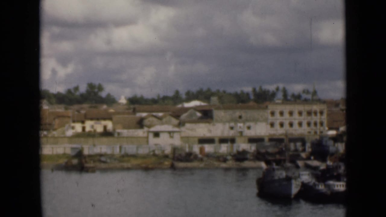 Colombo shoreline in archival 8mm footage, showing waterfront buildings, trees, moored boats, and a cloudy sky.
