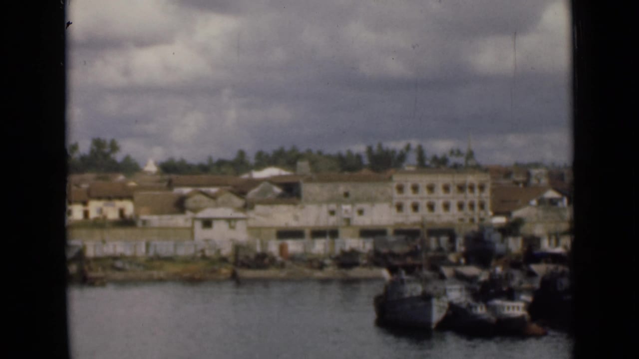 1940s Colombo waterfront scene with boats near shore, low-rise buildings, trees, and visible home-movie film grain.