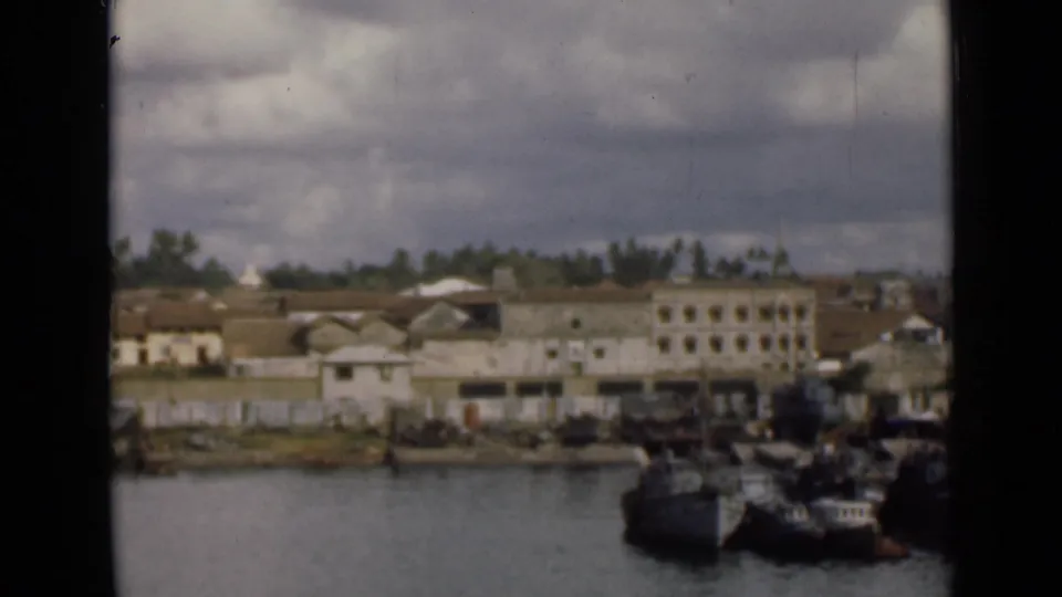 1940s Colombo waterfront scene with boats near shore, low-rise buildings, trees, and visible home-movie film grain.