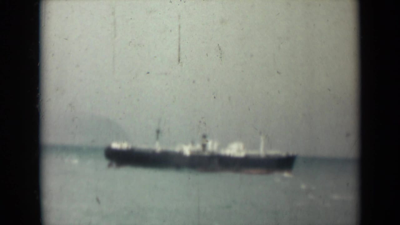 1946 India 8mm frame showing a dark-hulled cargo ship far offshore against calm water and a hazy sky.