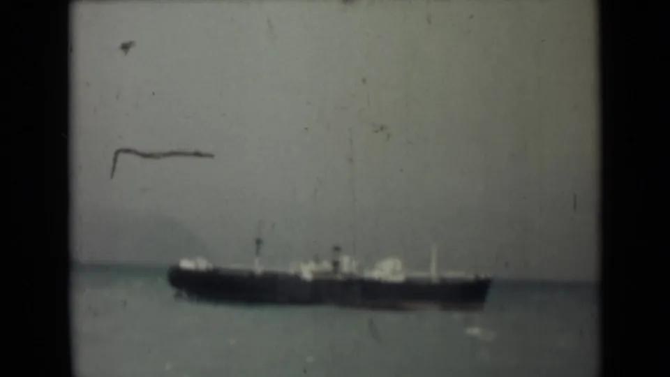 Soft 8mm view of a cargo ship offshore in India, with a dark hull, pale structures, calm sea, and visible film marks.