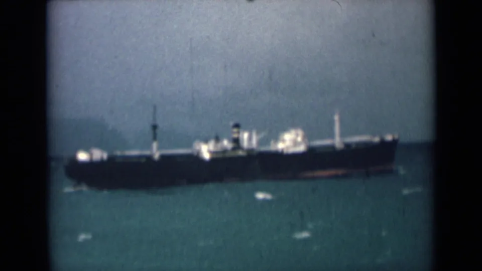 A large cargo vessel appears in profile on open water, with soft 8mm color and scratches visible across the frame.