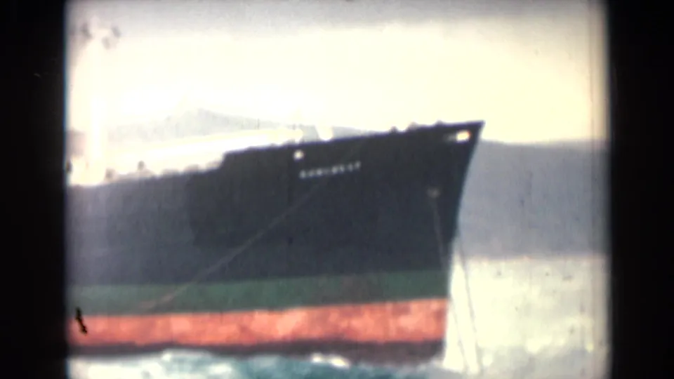 Closer 1946 8mm view of a cargo ship bow near coastal water, with overexposed sky and a red band along the lower hull.