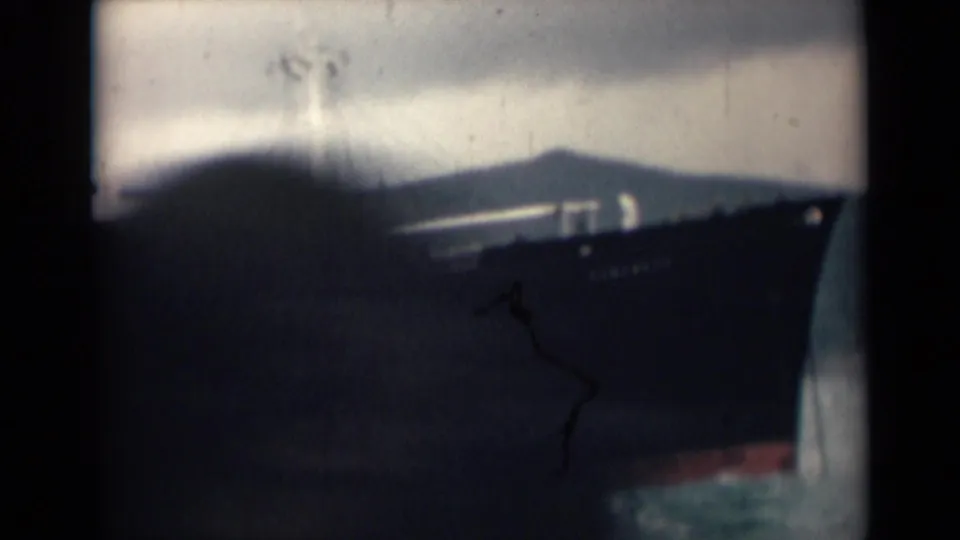 A dark ship bow fills the right side of the frame, with hazy hills and open water behind it in faded 8mm color.