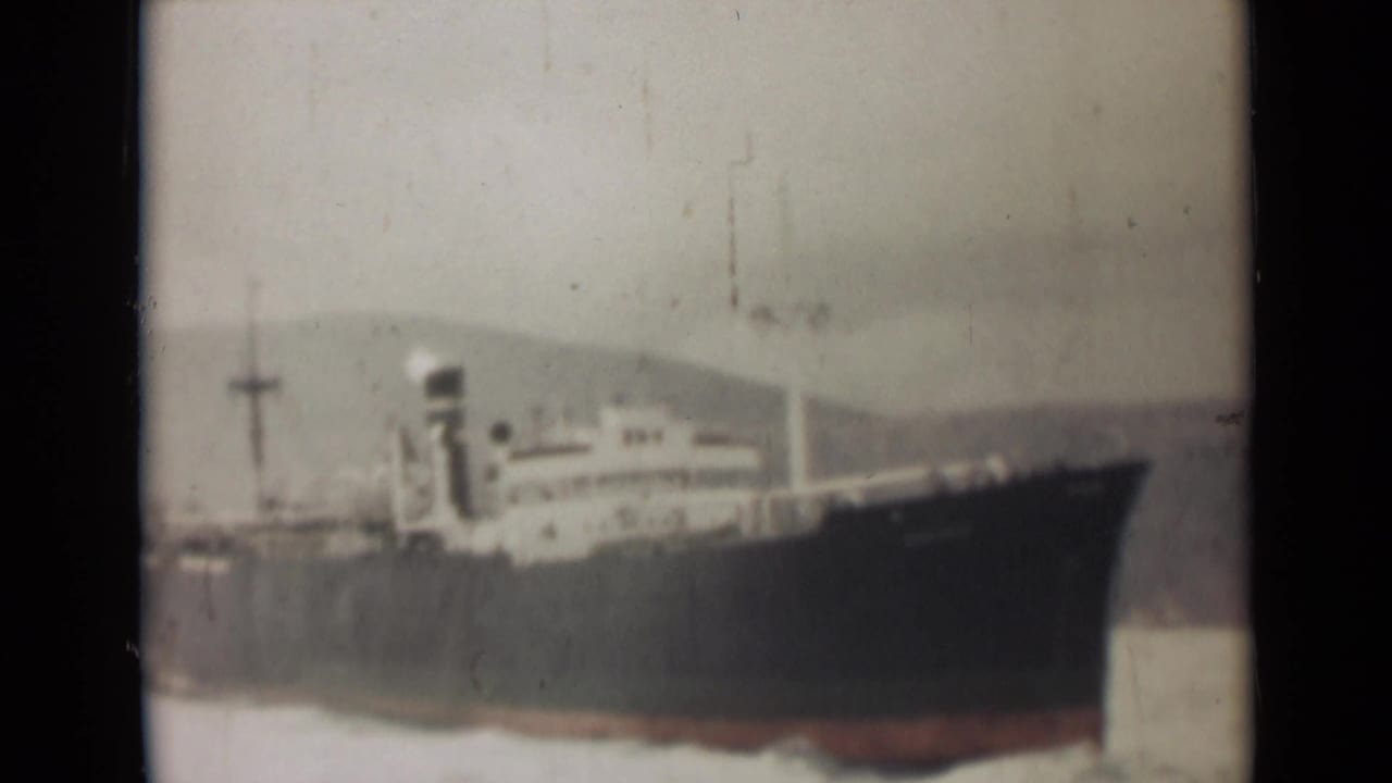 Large dark ship on calm water with hazy hills behind it.