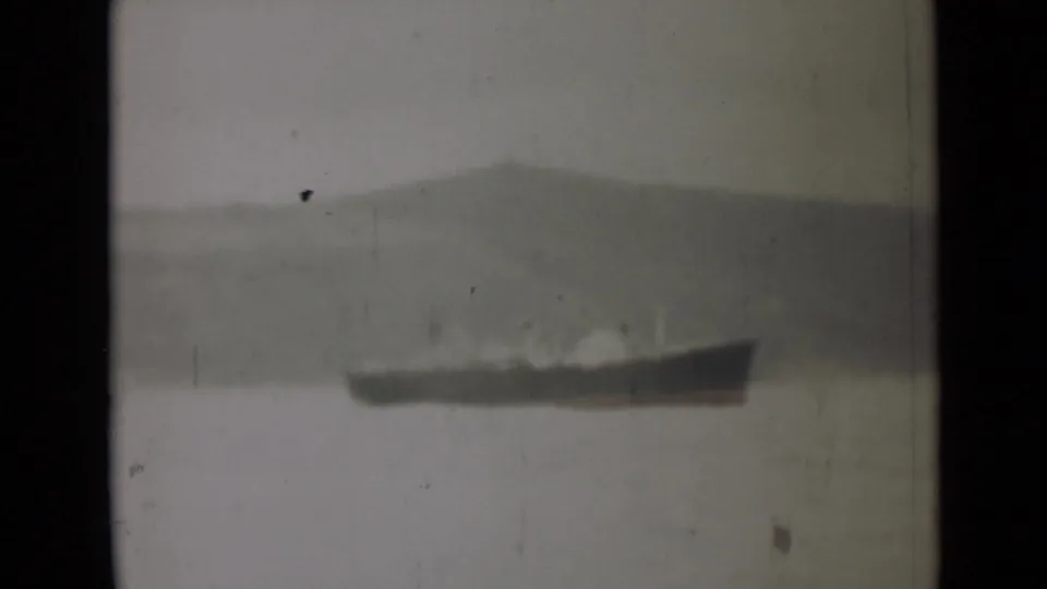 1946 India 8mm frame with a distant dark vessel crossing calm water beneath a soft, hazy hillside.