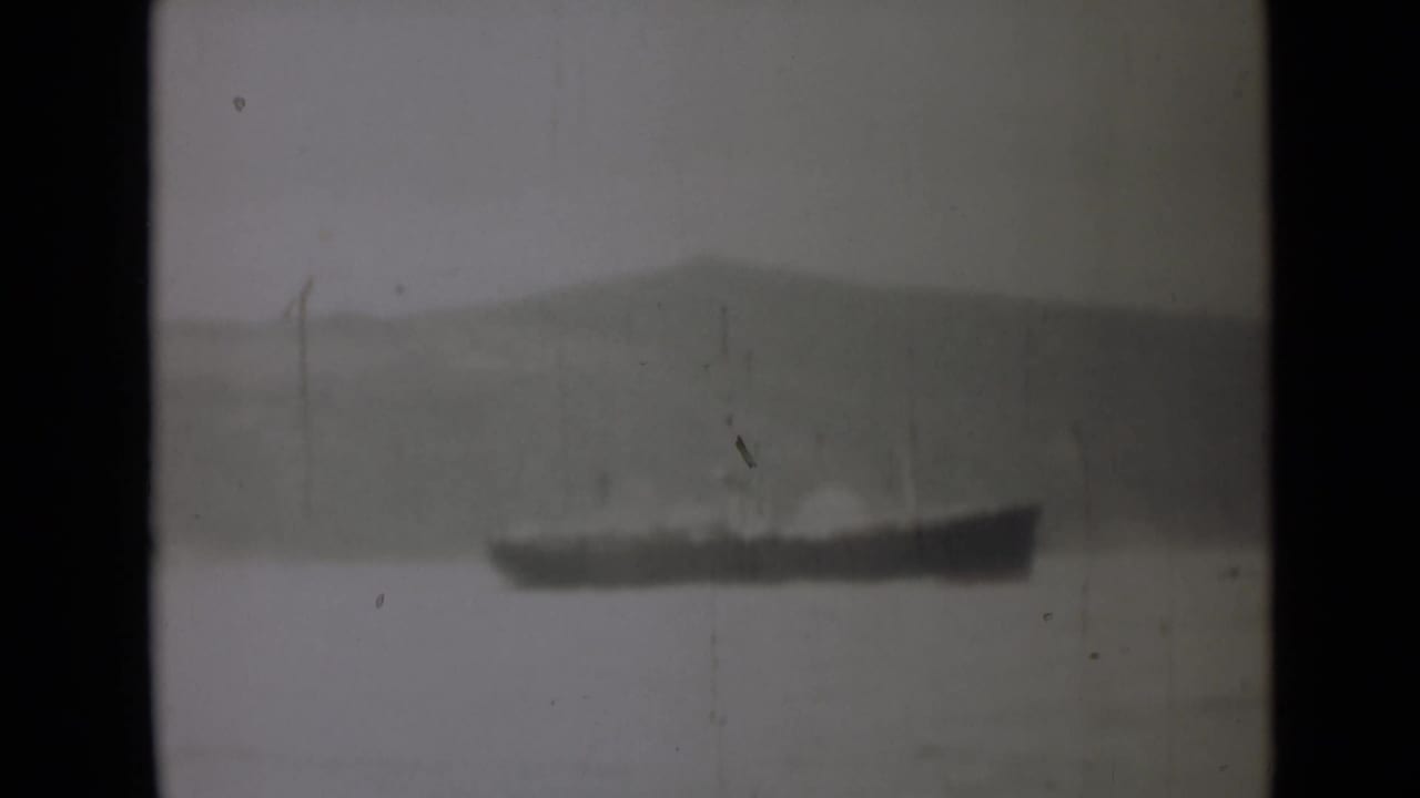 1946 India home-movie frame showing a distant dark vessel or landform across water under a hazy sky.