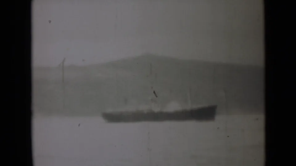 1946 India home-movie frame showing a distant dark vessel or landform across water under a hazy sky.
