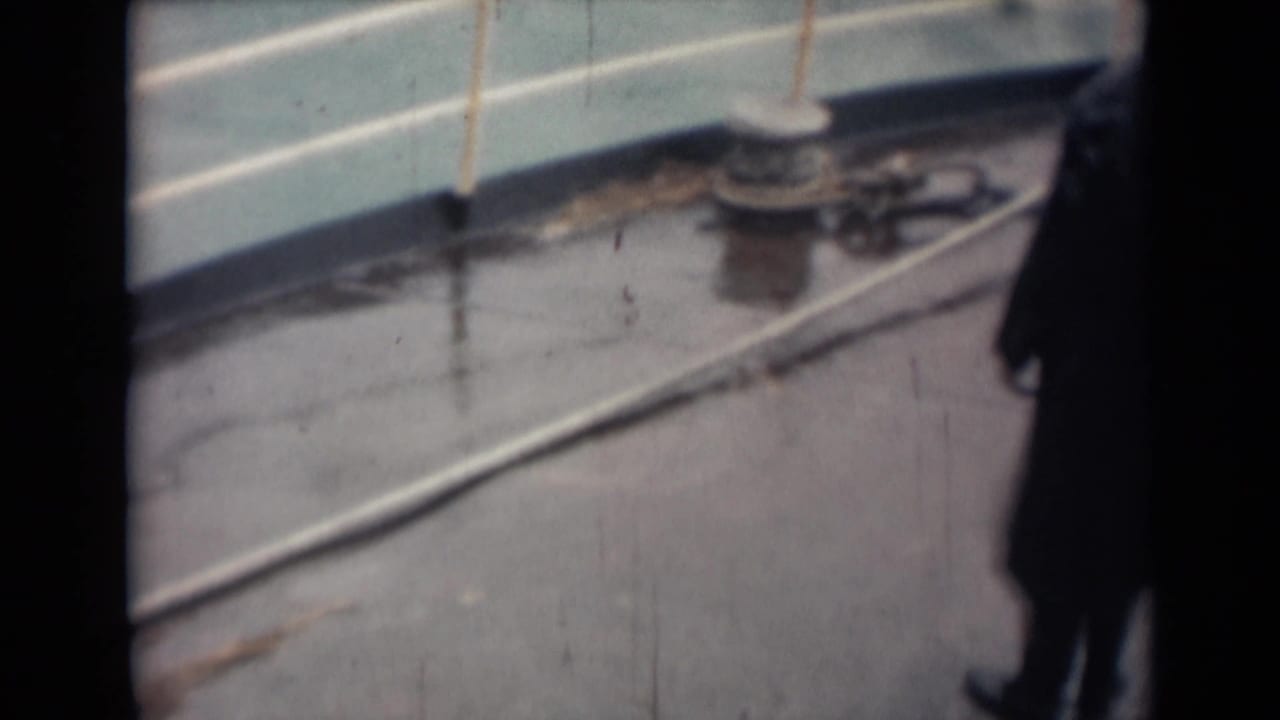 1946 India 8mm frame showing a rain-slick boat deck with white railing, bollard, rope, and a partially visible man in a dark coat.
