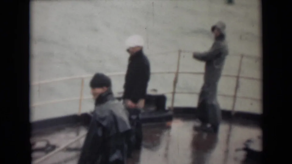 1946 India home-movie frame showing three men in dark coats and hats on a rain-wet vessel deck with railing and water in the background.