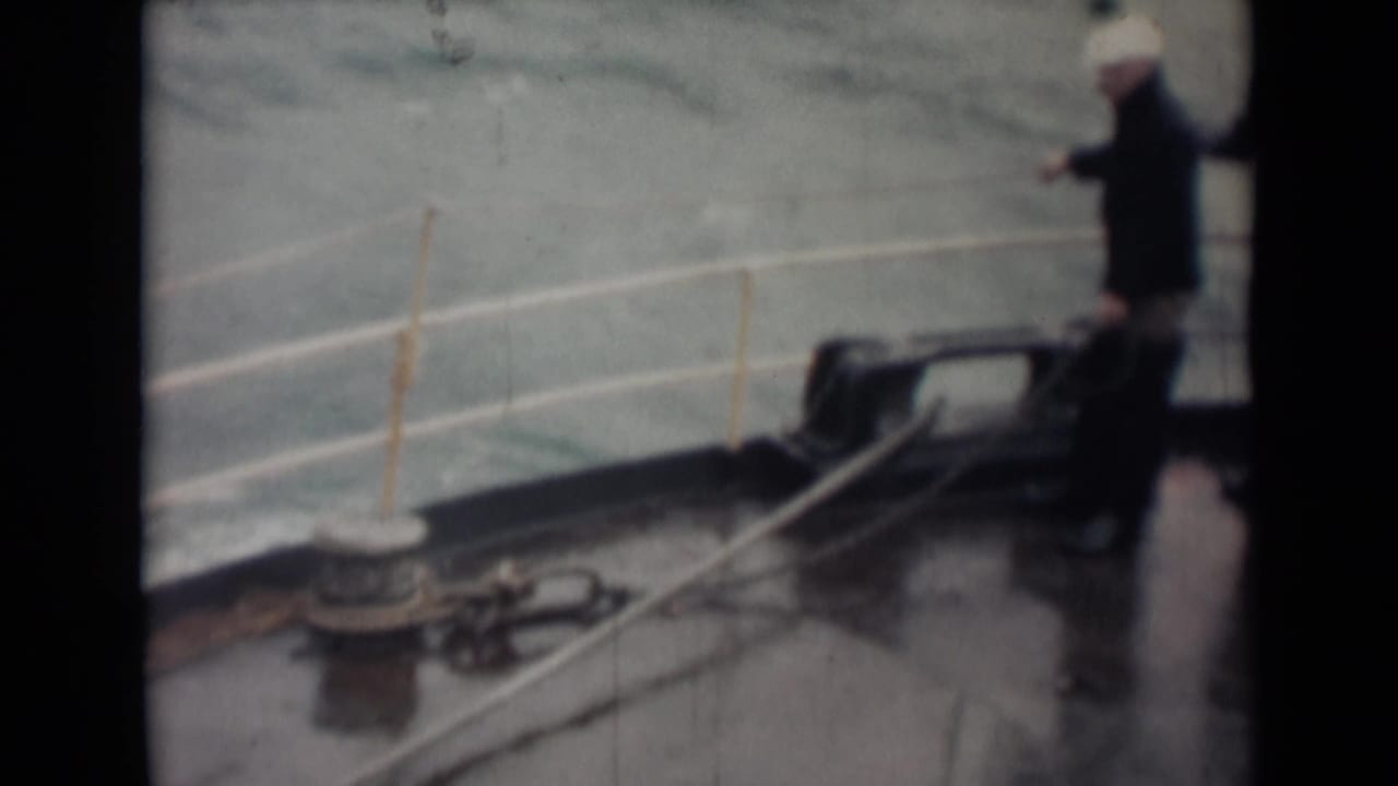 Two men in coats stand on a wet ship deck beside railings and deck hardware as rough water fills the background.