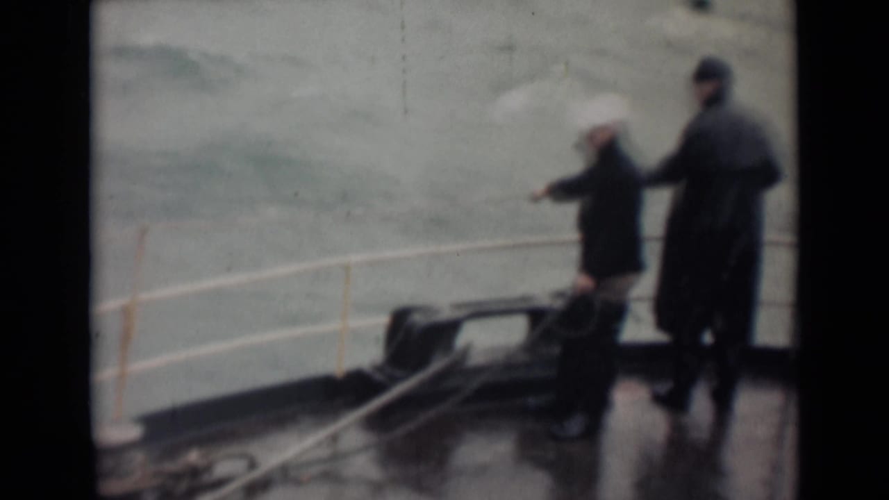 A man with a pale umbrella or hat-like covering stands near a railing while another man in a coat watches from the deck.