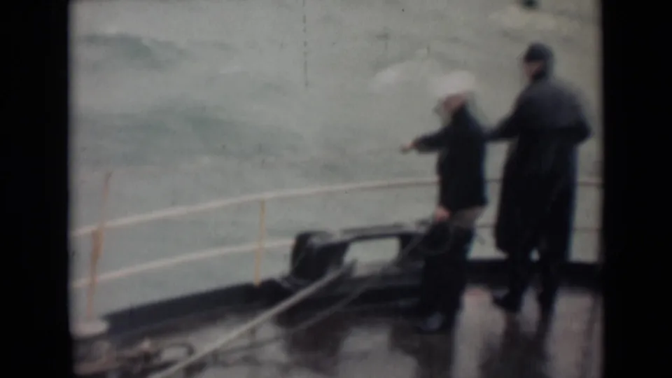 On a wet ship deck, two men in heavy outerwear stand near railings with choppy water behind them in 1946 India maritime footage per metadata.