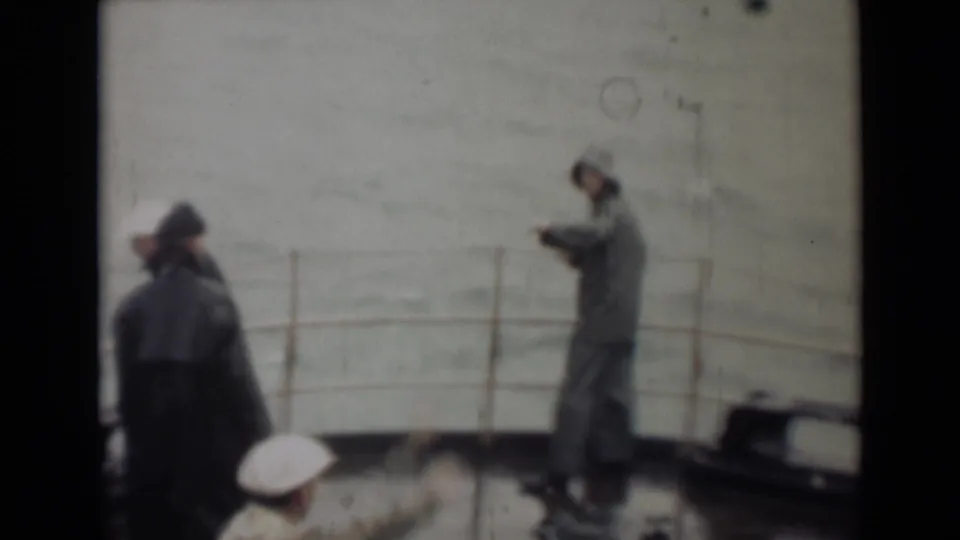 A deck-level 8mm view shows coated men near the ship railing, one handling or checking an object while rough sea stretches behind.
