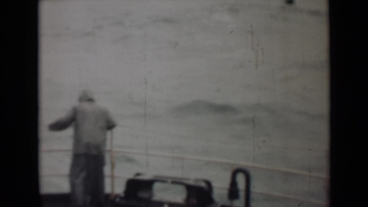 Man in a hooded coat stands by a boat railing facing rough water.