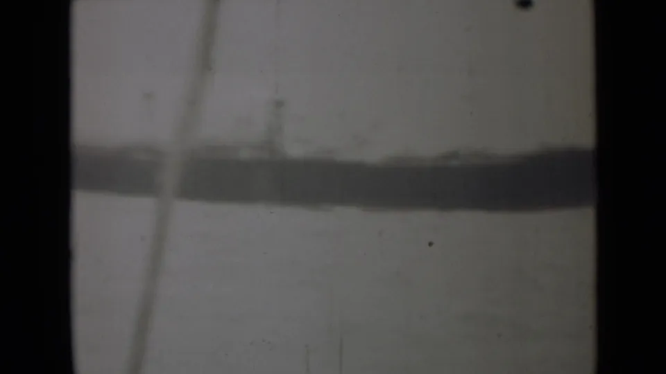 The frame shows a long dark vessel-like shape across pale water, partly obscured by soft focus and film wear.