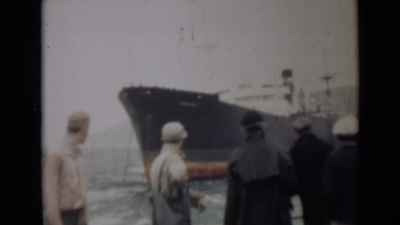 Several men stand near water looking toward a large dark ship with a red lower hull.
