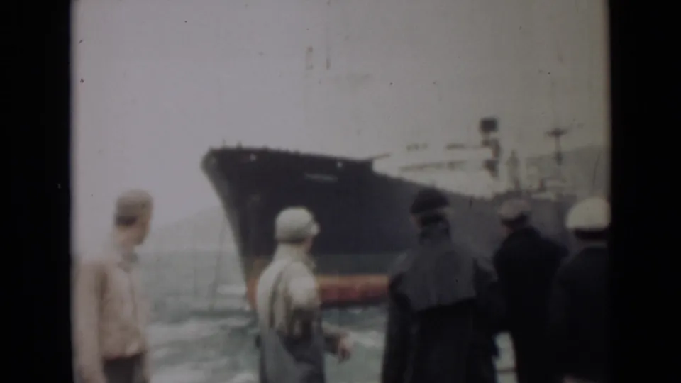 Men in period clothing stand by the water in India, 1946, watching a large grey ship with a red waterline under cloudy sky.