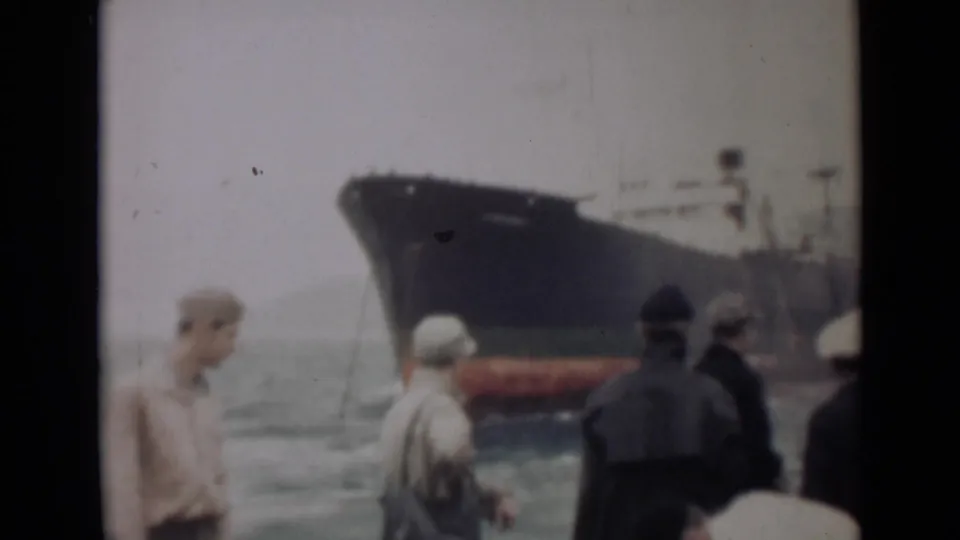A handheld 1946 India frame shows men in caps and coats facing a large ship, with water and hazy sky behind them.