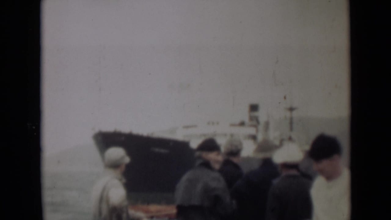 1946 India 8mm frame showing people gathered by choppy water as a large dark-hulled ship sits in the background.