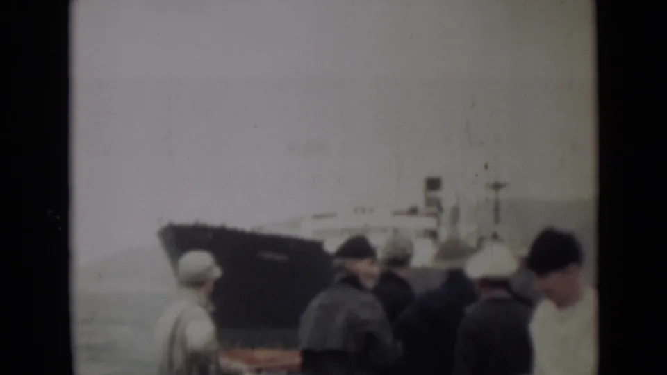 1946 India 8mm frame showing people gathered by choppy water as a large dark-hulled ship sits in the background.