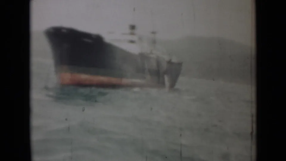 1946 India 8mm view of a large cargo vessel at sea, with a dark hull, red waterline, and hazy land beyond.