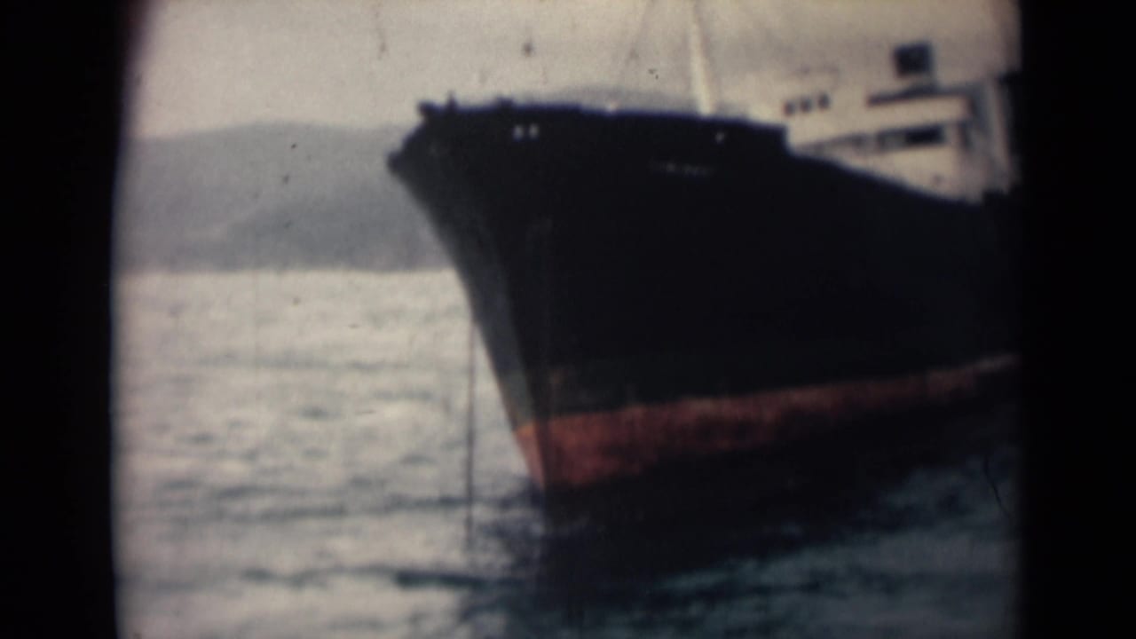 A large dark-hulled cargo or fishing vessel appears at sea in 1946 India footage, with distant shoreline or hills behind it.