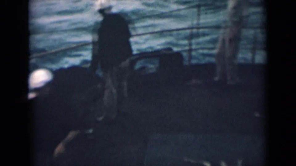 1946 India home-movie frame showing a dim ship deck, a sailor in a white cap, railings, and water behind him.