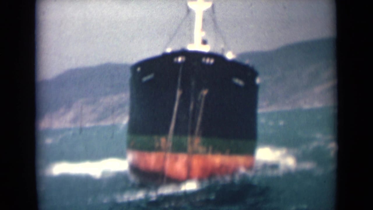 1946 India 8mm frame showing the cargo ship head-on, its black and red hull pushing through rough water near a hilly shoreline.