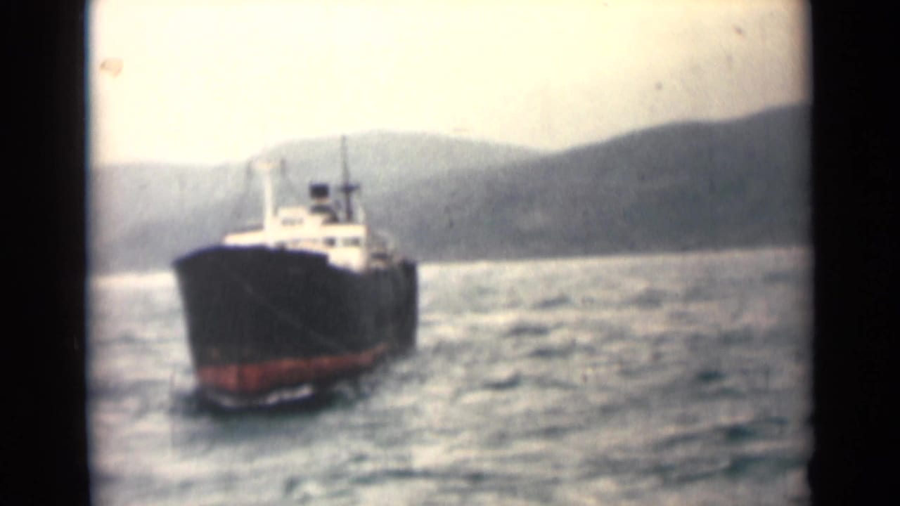 Cargo ship at sea viewed from the front, with hills and rough water behind it.