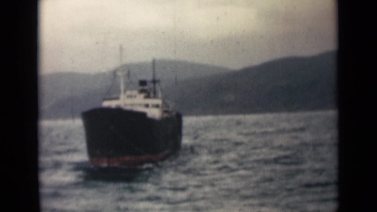 Home-movie view of the ship farther back on the water, with the coast and hills visible through muted 8mm color.