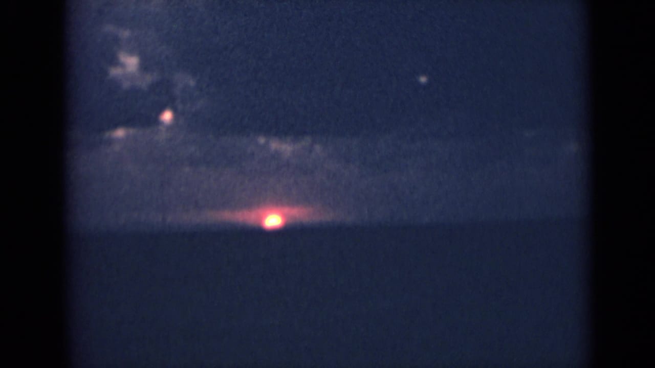 Archival 8mm sunset over India, with a red sun near the horizon below dark cloud cover.