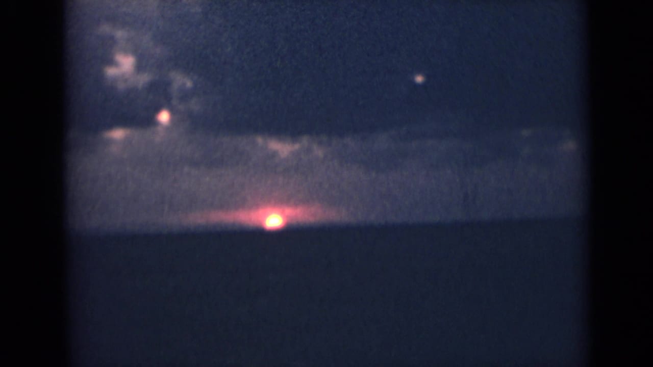 1940s India home-movie frame with a dark horizon, clouded sky, and a small bright glow near the lower center of the scene.