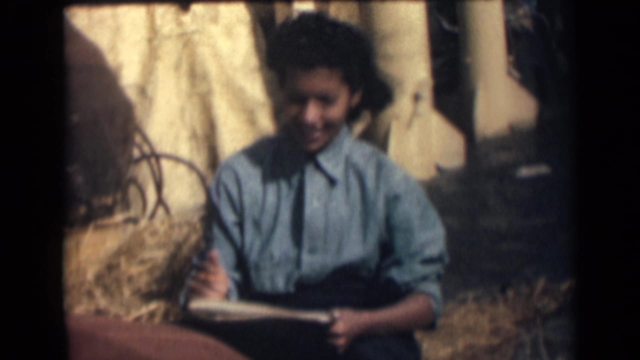 1946 India home-movie frame showing a young woman in a blue shirt seated on hay or straw with a notebook in a rural outdoor setting.